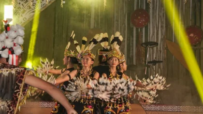 Penari perempuan berbusana adat Dayak hitam bermotif manik-manik dan hiasan kepala bulu sedang menari memegang properti bulu di atas panggung kayu. Panggung disorot lampu kuning dengan latar dinding bambu berhias perisai tradisional.