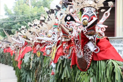Peserta Hudoq berbaris mengenakan topeng bermotif merah, putih, dan hitam, hiasan bulu, kain merah, serta balutan daun hijau.