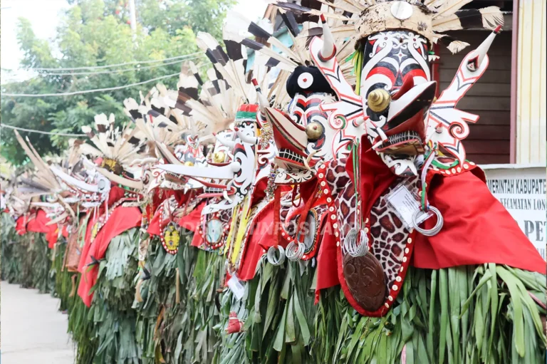 Peserta Hudoq berbaris mengenakan topeng bermotif merah, putih, dan hitam, hiasan bulu, kain merah, serta balutan daun hijau.